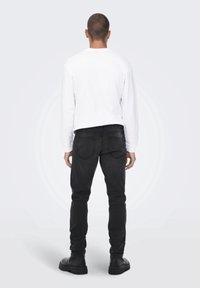 Camicia bianca a maniche lunghe, jeans in denim nero e stivali neri. I jeans hanno tasche posteriori e una vestibilità aderente. Sfondo neutro.