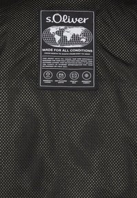 s.Oliver apģērbu etiķete ar pasaules karti un tekstu, kas izceļ jaku piemērotību visiem laikapstākļiem un ērtības funkcijas.