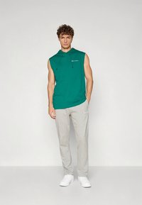 Roheline varrukateta kapuutsiga sviiter reguleeritavate nööridega, kombineeritud helehallide spordipükstega ja valgete tossudega. Kapuutsil on lihtne logo.