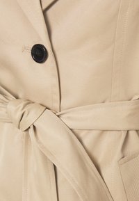 Beżowy płaszcz trench z czarnym guzikiem, przewiązywaniem w talii i delikatną, ukośną teksturą na materiale. Zawiera boczne kieszenie dla dodatkowego akcentu.