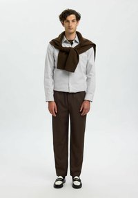 Gestreept overhemd, bruine wollen trui over de schouders gedrapeerd, bruine broek en bicolor schoenen. Neutrale achtergrond.