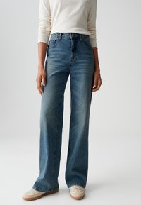 Light blue denim wide-leg jeans with faded sections, five pockets, and a classic button closure. Paired with beige sneakers.