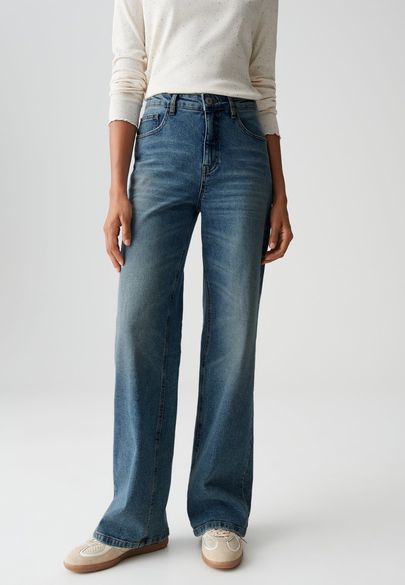 Light blue denim wide-leg jeans with faded sections, five pockets, and a classic button closure. Paired with beige sneakers.