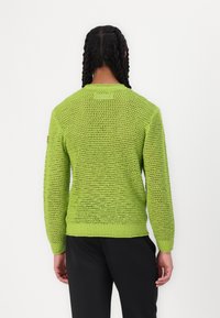 Pullover verde-lima tricotado, com um padrão de ponto aberto, punhos e bainha canelados, e um decote redondo. Usado com calças pretas.