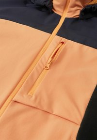 Gros plan sur l'avant d'une veste pêche et noire avec une fermeture éclair centrale et une poche poitrine verticale zippée.