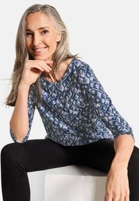 Mehrfarbige Bluse mit geometrischem Muster in Blau, Lila und Weiß. Sie hat einen Rundhalsausschnitt und Dreiviertelärmel. Figurbetonter Schnitt.
