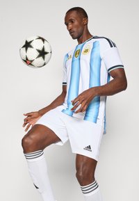 Atleet in Argentijns voetbaltenue beheerst een witte bal met zwarte sterren, zich voorbereidend om te trappen of te jongleren tegen een effen achtergrond.