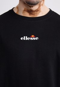 Camiseta de algodón negra con un logo blanco de "ellesse" y un diseño de acento en rojo y naranja por encima del texto. Cuello redondo, mangas cortas.