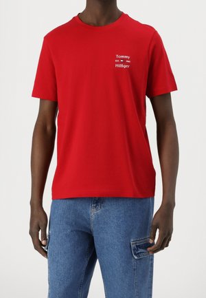 Mężczyzna ubrany w czerwoną koszulkę Tommy Hilfiger z logo na piersi oraz niebieskie dżinsy z boczną kieszenią.