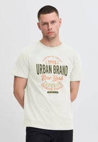 Svetlozeleno bombažno majico z grafičnim tiskom v oranžni in zeleni barvi, ki vsebuje besedilo "URBAN BRAND New York" ter okroglo ovratnico.