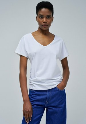 Camiseta de algodón blanca con escote en V y mangas cortas, con un trim texturizado en los dobladillos de las mangas. Combinada con pantalones de mezclilla azul.