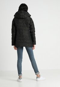Svart puffjacka med huva, med en quiltad design. Modellen bär ljusblå jeans och vita sneakers med gröna detaljer.