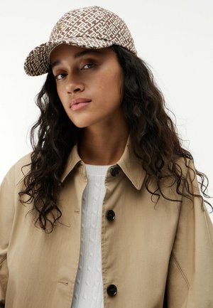 Vrouw met lang krullend haar die een bruin-witte pet met patroon draagt, een beige jas met knopen en een witte gebreide trui tegen een effen achtergrond.