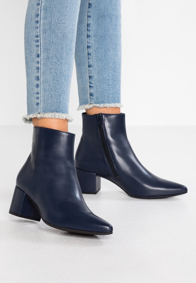 Personne portant un jean bleu clair à ourlet effiloché et des bottines en cuir bleu marine à talons carrés avec fermetures éclair latérales.