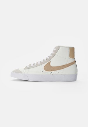 Białe sneakersy za kostkę z beżowymi zamszowymi akcentami, beżowym logo, białymi sznurowadłami i białą podeszwą na jednolitym tle.