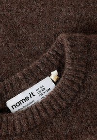 Braun gestrickter Stoff mit sichtbarem weißen Etikett, das den Markennamen, die Größenangaben und den Herstellungsort zeigt.