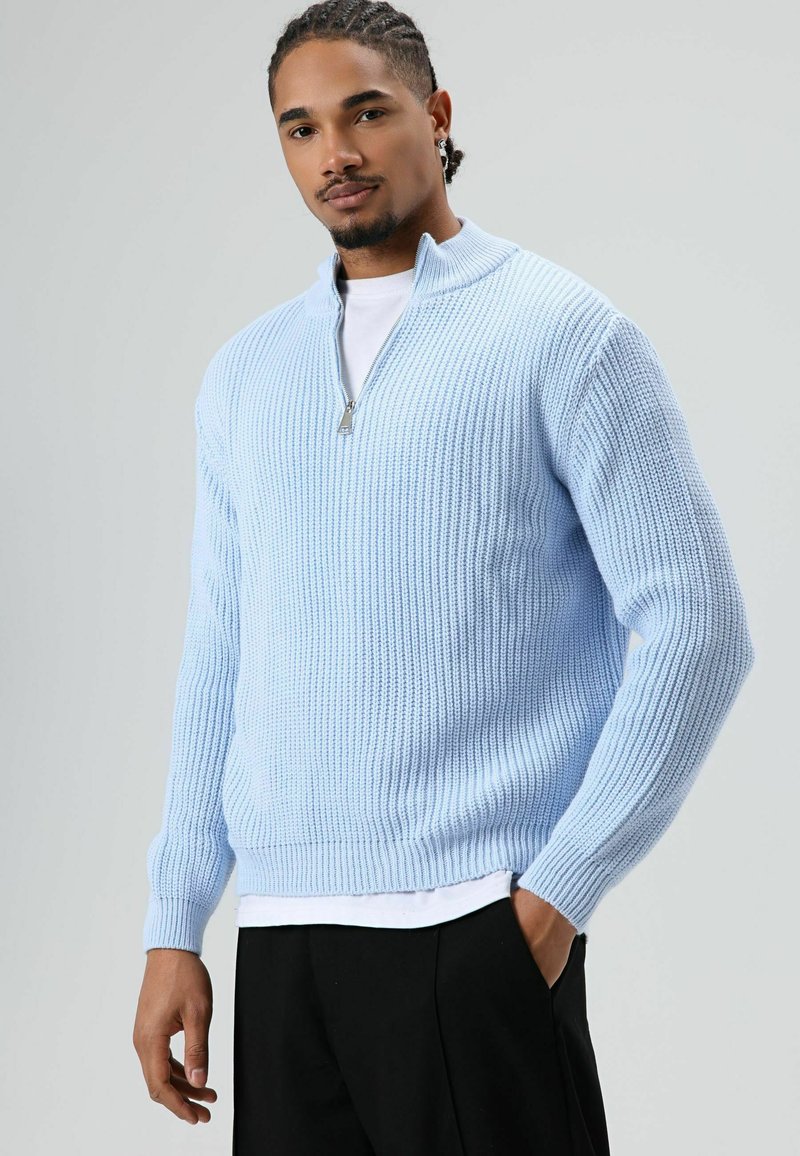 Homme aux cheveux tressés portant un pull en tricot bleu clair à demi-fermeture éclair, un t-shirt blanc et un pantalon noir, debout avec une main dans la poche.