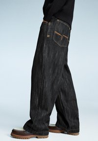 Black wide-leg jeans made of denim, featuring contrasting orange stitching, a back pocket design, and paired with brown shoes.
