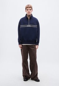 Μπλε ναυτικό πουλόβερ με γιακά με μερικό φερμουάρ, διακοσμημένο με λευκή οριζόντια ρίγα και το λογότυπο 'Calvin Klein'. Συνδυάζεται με φαρδιά καφέ παντελόνια.