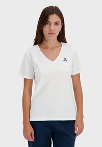 Bílý krátký rukáv tričko s výstřihem do V a vyšívaným logem na hrudi. Měkká bavlněná látka, volný střih, v kombinaci s námořnickými kalhotami.