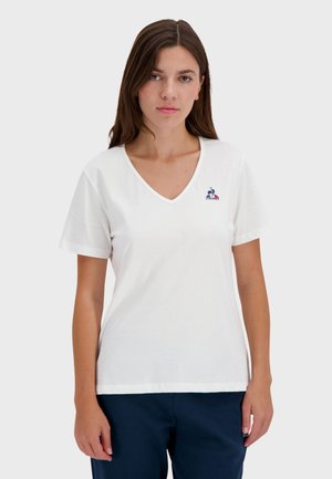 White short-sleeve T-shirt with a V-neck and embroidered logo on the chest. Soft cotton fabric, relaxed fit, paired with navy pants.