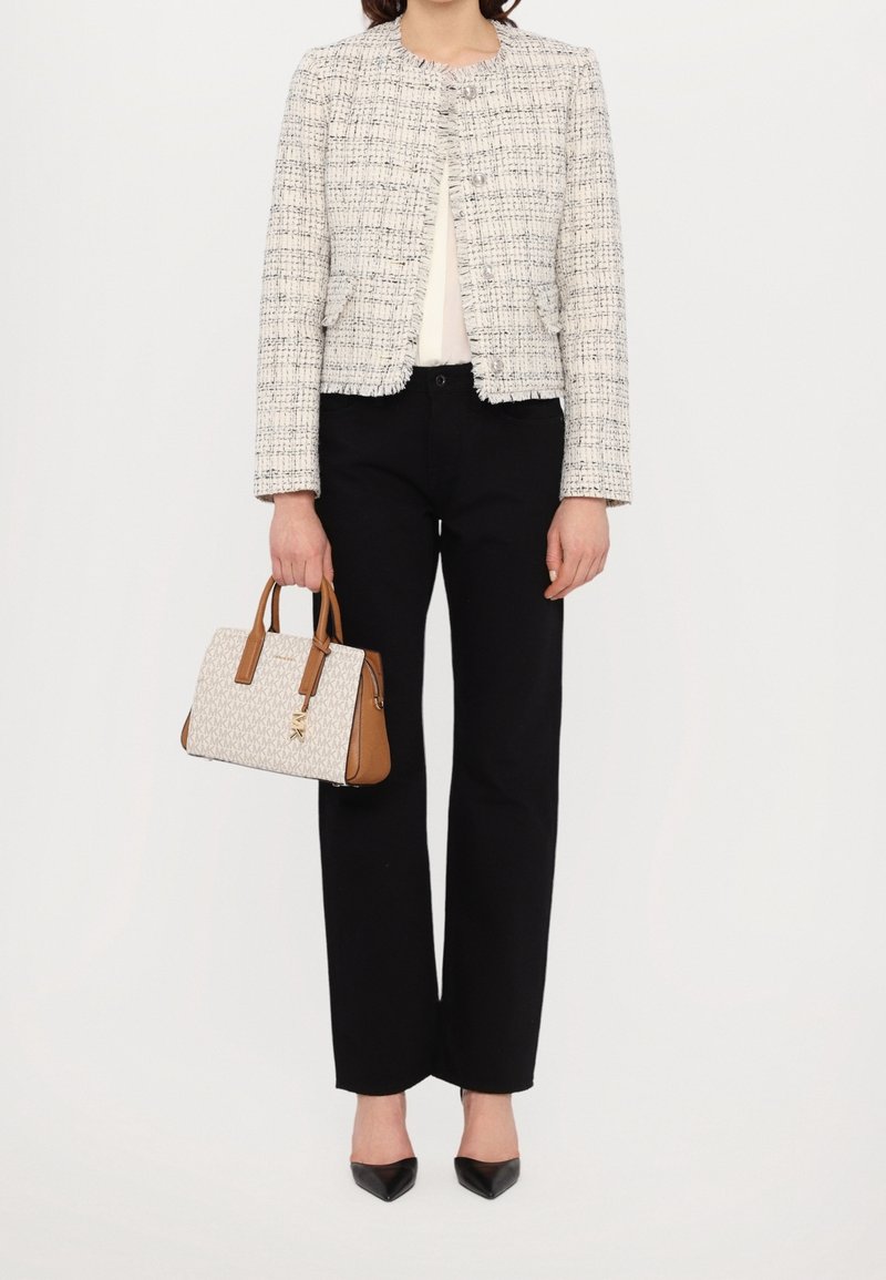 Personne portant une veste blanche texturée, un pantalon noir, des chaussures noires pointues, et tenant un sac à main blanc et marron avec un motif de logo.