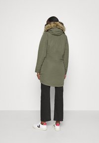 Olivgrön parka-jacka med huva av konstpelts, med långa ärmar och avskuren midja. I kombination med svarta byxor och vita sneakers.