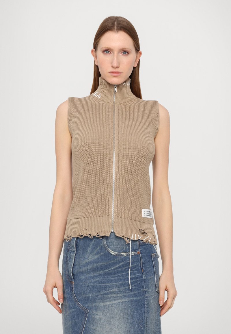 Beige knitted sleeveless top with a high collar and zip closure, featuring a frayed hem and label detail at the bottom.