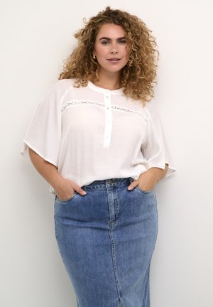 Bela bluzica s gumbi in čipkastimi detajli, v kombinaciji z modrim denim krilom z visokim pasom in sprednjim razporkom, oba delujeta sproščeno in prilegajoče.