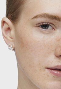 Srebrne kolczyki w kształcie kółek prezentowane na uchu modelki, z gładkim, wypolerowanym wykończeniem i prostym, zaokrąglonym kształtem. Skóra ma naturalną, piegowatą teksturę.