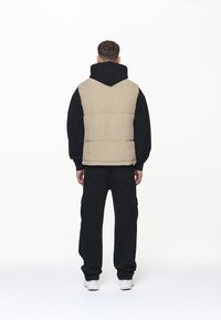 Béžová manšestrová prošívaná vesta v kombinaci s černou mikinou s kapucí, černými nákladními kalhotami a bílými teniskami; zobrazeno zezadu na jednoduchém pozadí.