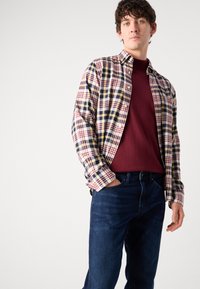 Checked shirt in white, red, blue, and yellow; long sleeves rolled up; worn over a burgundy sweater; paired with dark blue jeans.