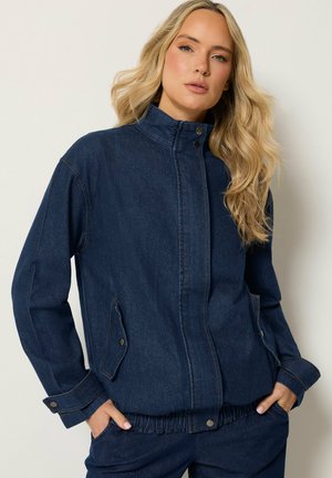 Frau mit langen blonden Haaren, die eine dunkelblaue Jeansjacke und passende Hose trägt und mit den Händen in den Taschen vor einem einfarbigen Hintergrund steht.