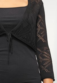 Black cropped cardigan with openwork design and long sleeves, featuring a tie closure at the front and a textured knit pattern.