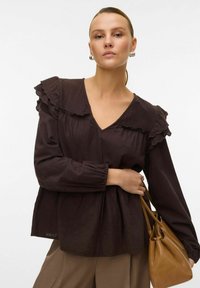 Blouse marron foncé avec col et manches à volants, décolleté en V, taille froncée et tissu texturé, associée à un sac à main beige.