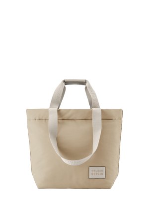 Beige Tragetasche mit zwei weißen Stoffgriffen und einem kleinen rechteckigen Aufnäher mit der Aufschrift Studio Berlin vorne unten rechts.