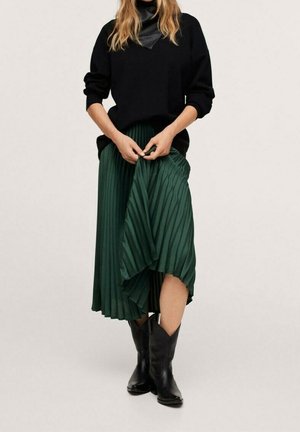 Femme portant un pull noir avec un détail en cuir, une jupe midi plissée verte et des bottes noires mi-mollet, debout devant un fond uni.