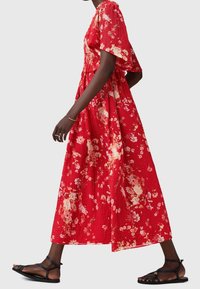 Femme en robe midi rouge fleurie à manches courtes marchant, portant des sandales plates noires à brides et peu de bijoux sur un fond uni.