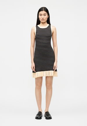 Ung kvinde iført en ærmeløs sort og beige stribet strikket kjole med plisseret kant og sorte loafers, stående foran en ensfarvet baggrund.