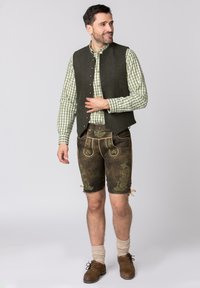 Traditioneel Beiers outfit met een olijfgroene wollen vest, groen geruit shirt, bruine leren shorts met geborduurde bloemenpatronen, en bruine schoenen.
