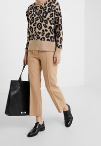 Persona con un suéter de estampado de leopardo, pantalones cortos beige, zapatos de cuero negros, sosteniendo un gran bolso tote negro con el logo blanco de MSGM.
