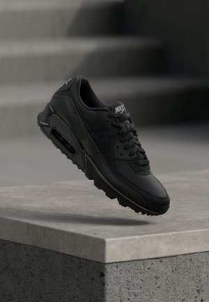La chaussure Nike Air Max 90 noire présente une combinaison de matériaux en cuir et en mesh, des lacets ronds, et une unité d'air visible sur la semelle.