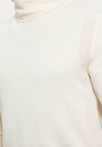 Cream turtleneck sweater in a soft knit fabric featuring ribbed texture and a subtle logo on the chest. Side has a patterned knit detail.