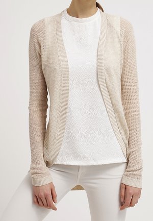 Femme portant un cardigan beige en maille ouvert sur le devant, par-dessus un haut blanc texturé et un pantalon blanc, debout les mains détendues le long du corps.