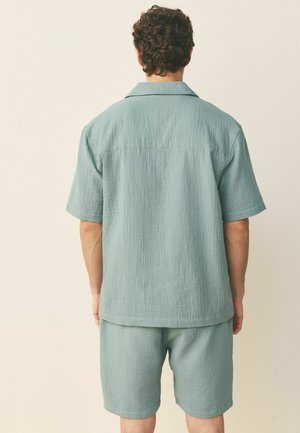 Man wearing light blue textured short-sleeve shirt and matching shorts, standing facing away against a plain off-white background.