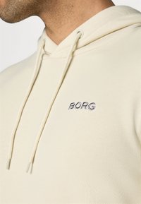 Hoddie beige clair avec une finition texturée, doté d'un cordon de serrage et d'un logo "BORG" brodé en bleu marine. Tissu doux, design décontracté.