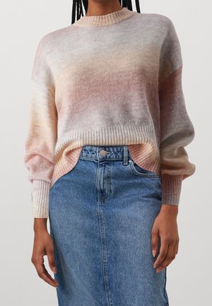 Žena v pastelovém ombre pleteném svetru, zastrčeném do vysoko pasové modré džínové sukně s předními kapsami a zapínáním na knoflíky.