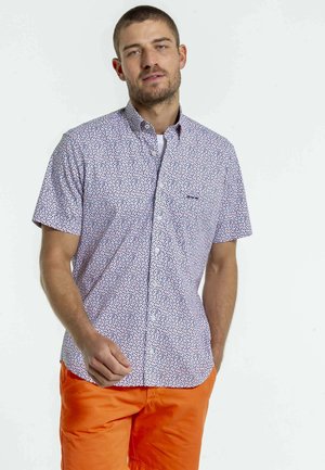 Chemise à manches courtes avec un fond blanc, un motif de pois bleu marine et rouge, et une poche sur la poitrine. Portée avec un short orange vif.
