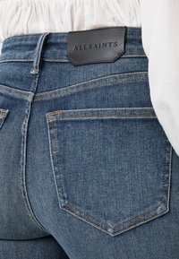 Denim jeans in medium wash met gestikte accenten, voorzien van een zwart lederen achterpatch met het label "ALLSAINTS" en twee achterzakken.