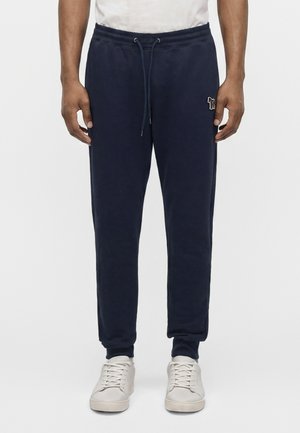 Homme portant un pantalon de jogging bleu marine avec taille à cordon et des baskets blanches, debout devant un fond uni.